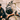 A black bicycle helmet with a built-in microphone and charging port hangs from the handlebars of a white bicycle. In the softly blurred background, a couple in black leather jackets sits together against a stone wall.